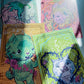Four illustrated art cards or prints arranged on a light surface, each with a whimsical animal character and decorative border. The visible designs include teal, pink, tan, and lime green backgrounds with colorful line art and floral details.