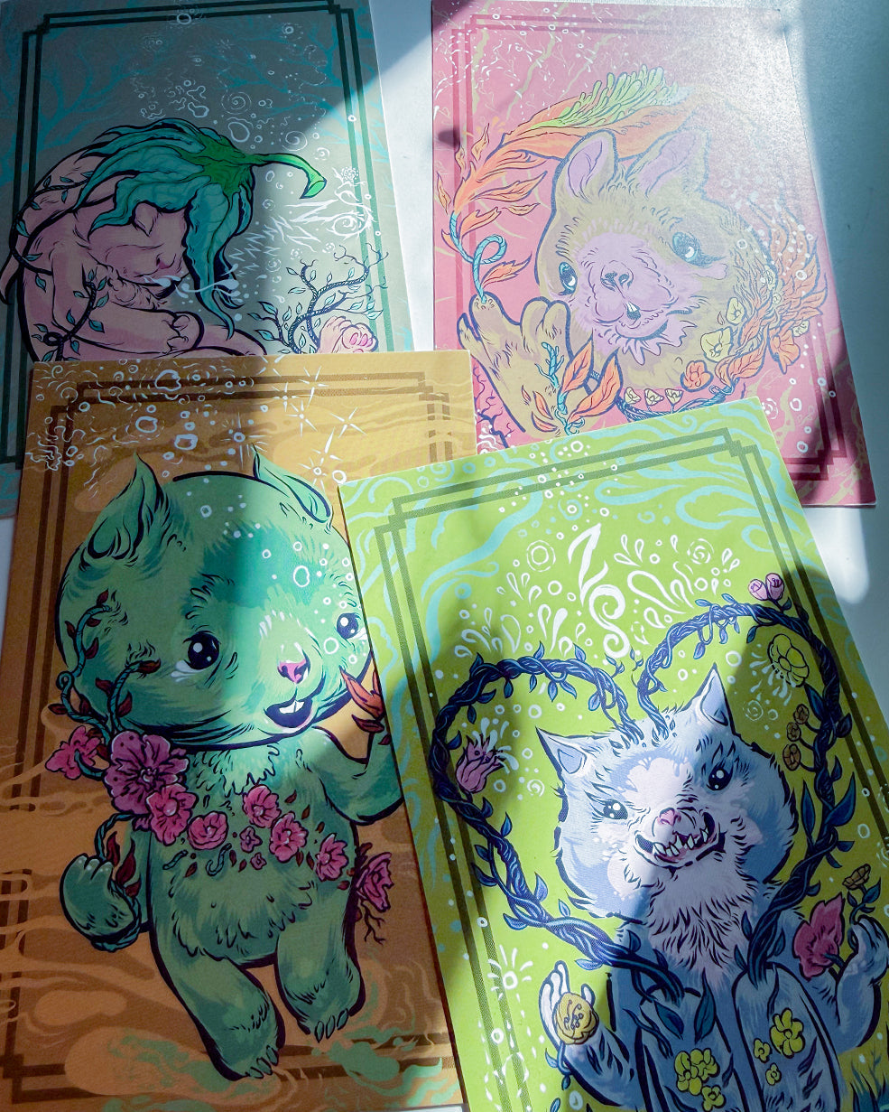 Four illustrated art cards or prints arranged on a light surface, each with a whimsical animal character and decorative border. The visible designs include teal, pink, tan, and lime green backgrounds with colorful line art and floral details.