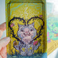 A hand holds a square illustrated card featuring a gray cat-like creature surrounded by dark vines and pink flowers on a yellow background. Another card is partially visible behind it on the right.