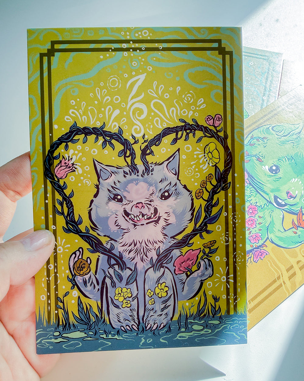 A hand holds a square illustrated card featuring a gray cat-like creature surrounded by dark vines and pink flowers on a yellow background. Another card is partially visible behind it on the right.