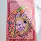 A square art print featuring a stylized cartoon alpaca in the center, surrounded by colorful abstract brushstrokes and decorative borders on a pink background, photographed against a white wall.