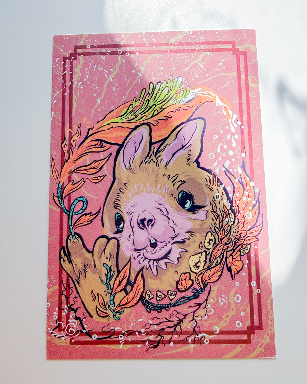 A square art print featuring a stylized cartoon alpaca in the center, surrounded by colorful abstract brushstrokes and decorative borders on a pink background, photographed against a white wall.