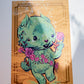 A square art print featuring a cute mint green cartoon bear holding pink flowers, set against a warm orange background with decorative border details.