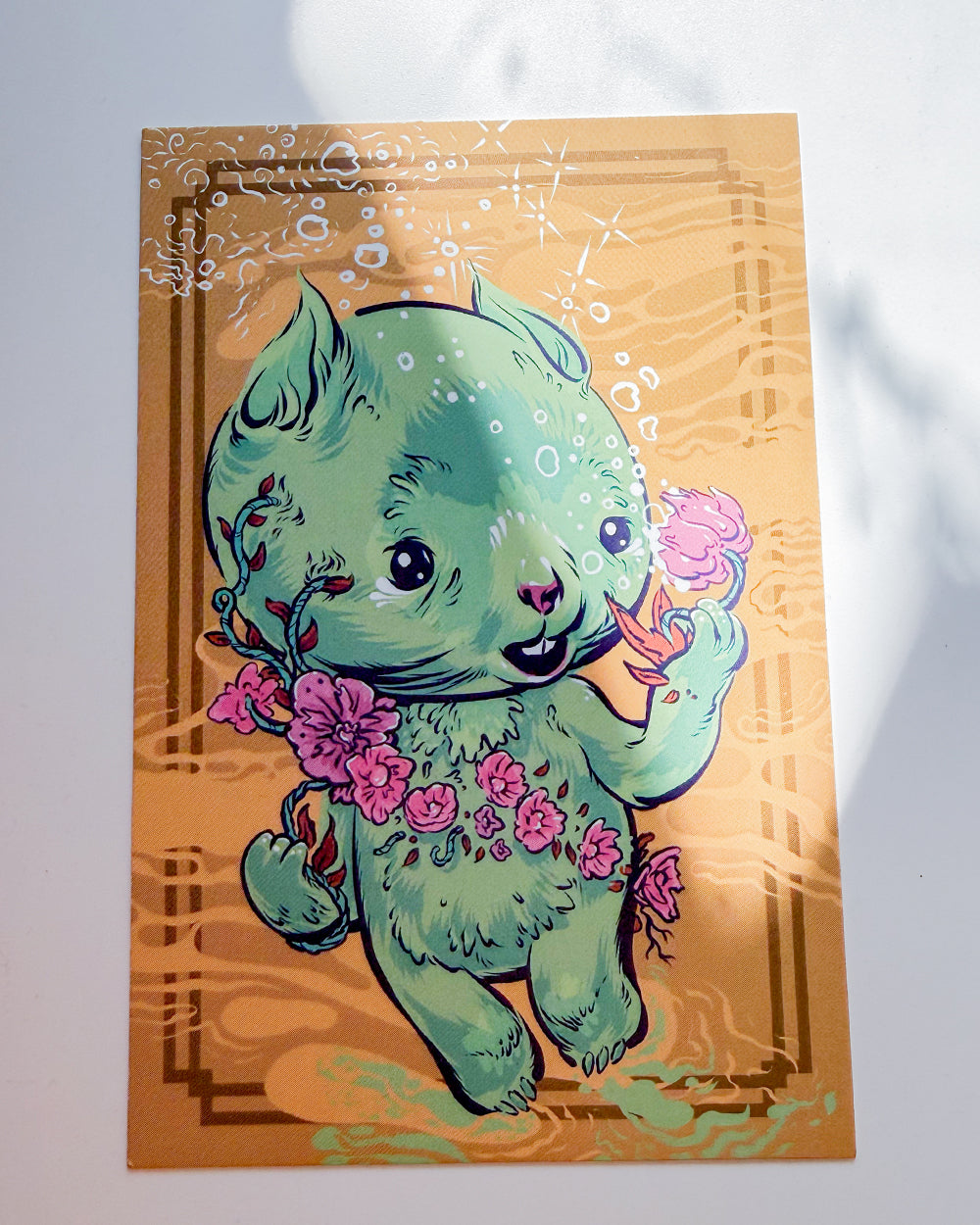 A square art print featuring a cute mint green cartoon bear holding pink flowers, set against a warm orange background with decorative border details.