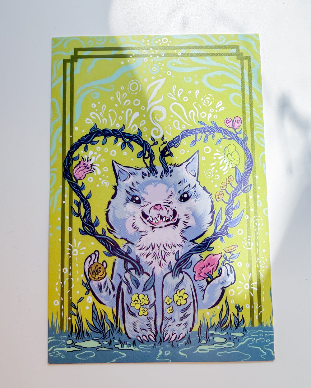 A square art print featuring a whimsical illustration of a blue-gray cat with a flower-patterned shirt, surrounded by vines and pink flowers on a bright yellow-green background, photographed against a white wall.