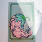 A square art print featuring a stylized illustration of a fetus with green leaf-like hair, set against a pale background with a decorative border, photographed on a white surface with soft shadows.