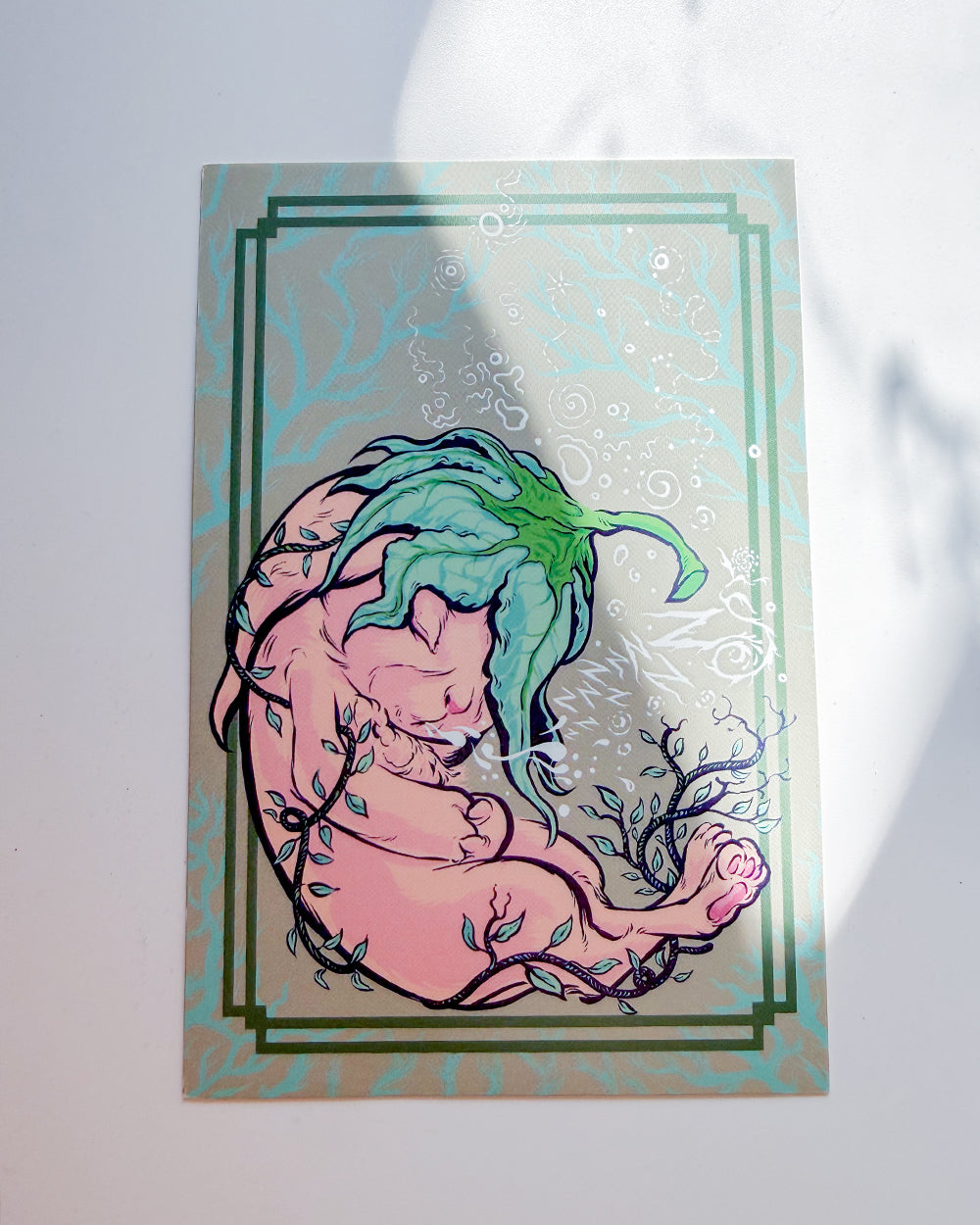 A square art print featuring a stylized illustration of a fetus with green leaf-like hair, set against a pale background with a decorative border, photographed on a white surface with soft shadows.