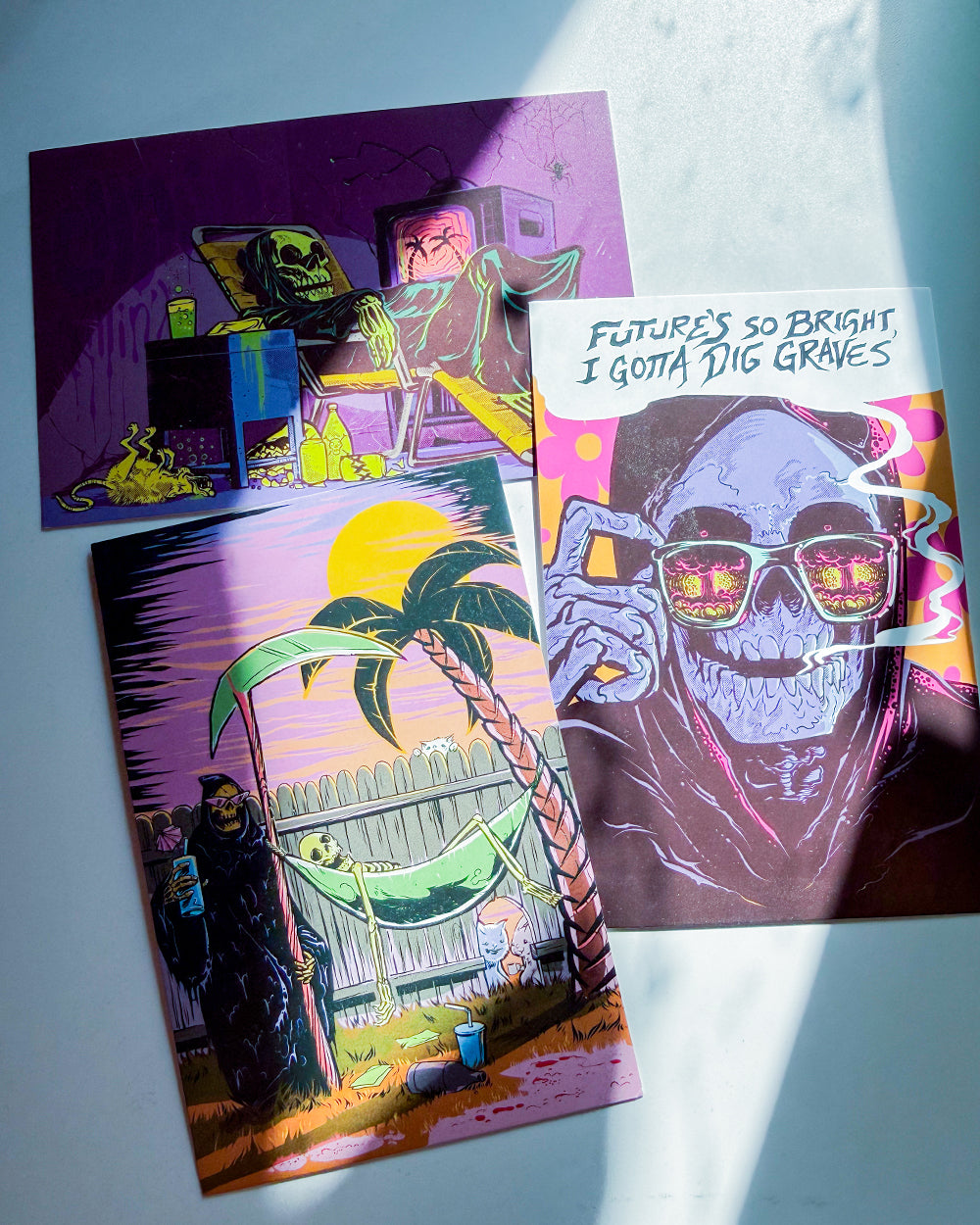 Three colorful art prints featuring surreal, horror-themed illustrations are arranged on a light blue surface with natural sunlight casting shadows. The prints depict a character in a black robe holding a cup next to a hammock, a character in a lab coat looking at a brain, and a skull wearing sunglasses with the text 'FUTURE'S SO BRIGHT, I GOTTA DIG GRAVES'.