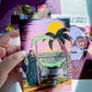 A hand holds a colorful illustrated art card featuring a surreal scene with a palm tree, a hammock, and two skeletal figures under a yellow sun. Other art cards are partially visible in the background on a white surface.