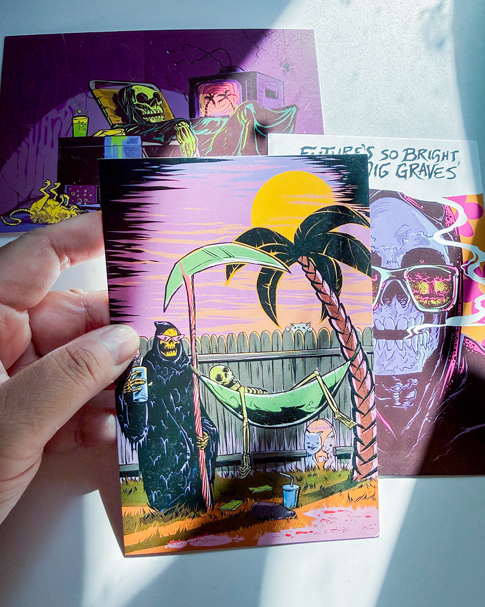 A hand holds a colorful illustrated art card featuring a surreal scene with a palm tree, a hammock, and two skeletal figures under a yellow sun. Other art cards are partially visible in the background on a white surface.