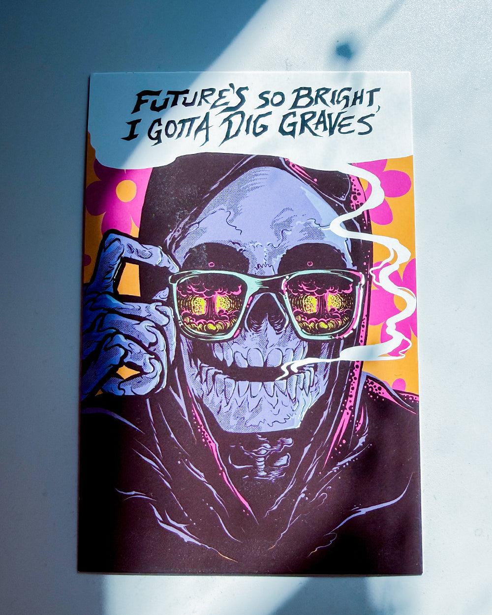 A rectangular art print or card featuring a stylized skull wearing sunglasses with colorful reflections, plus a hand holding a phone near the face. The top text reads, 'FUTURE'S SO BRIGHT I GOTA DIG GRAVES.