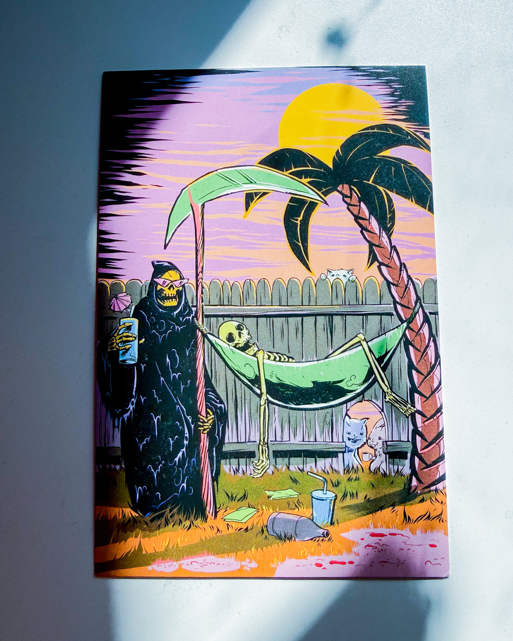 A colorful art print or card featuring a grim reaper holding a scythe next to a hammock with a skull, under a yellow sun and palm tree, photographed against a light gray background with shadows.