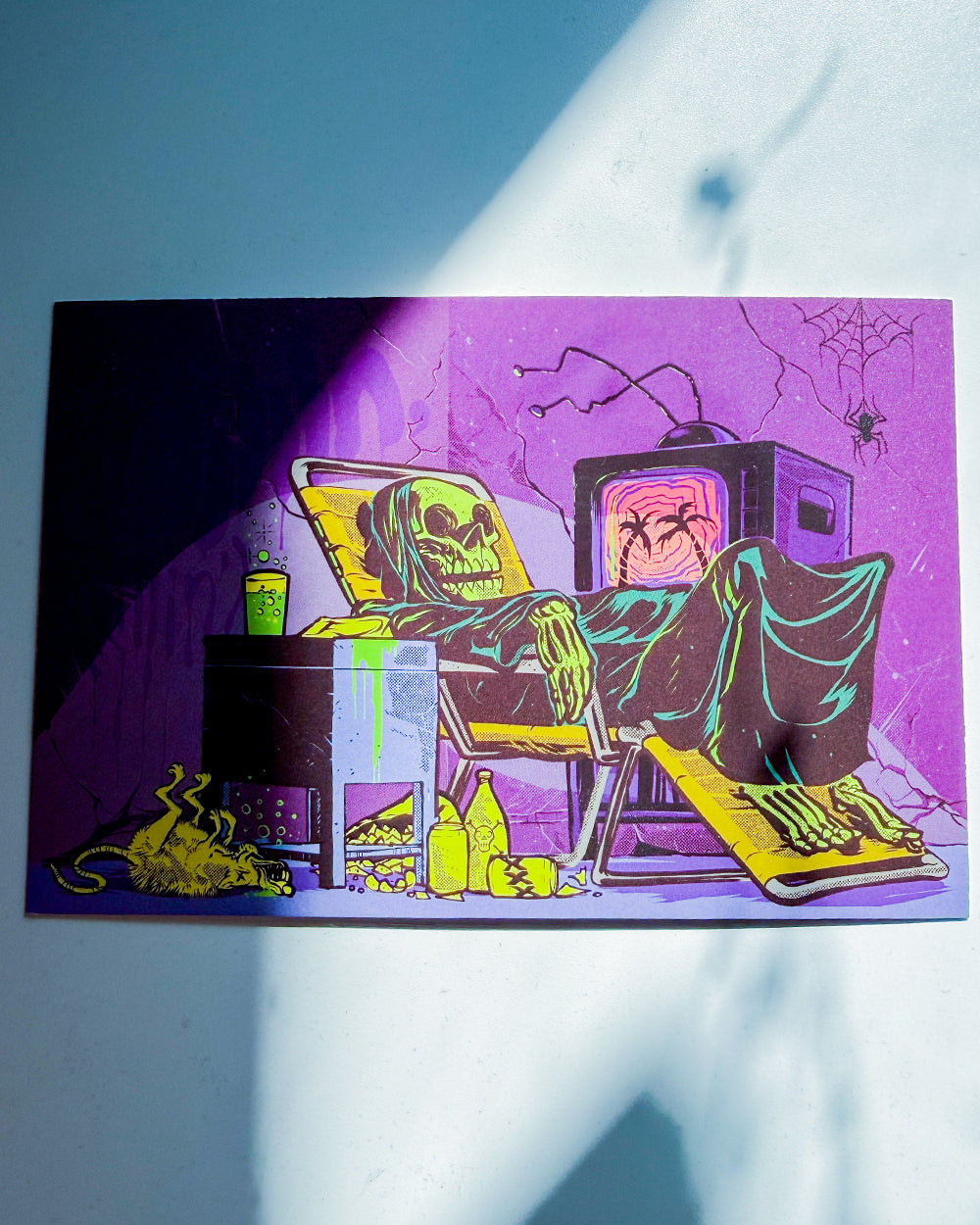 A square art print or card featuring a psychedelic horror illustration of a skeleton reclining in a chair next to a television, with neon purple, yellow, green, and black colors, photographed on a light surface with strong diagonal shadows.