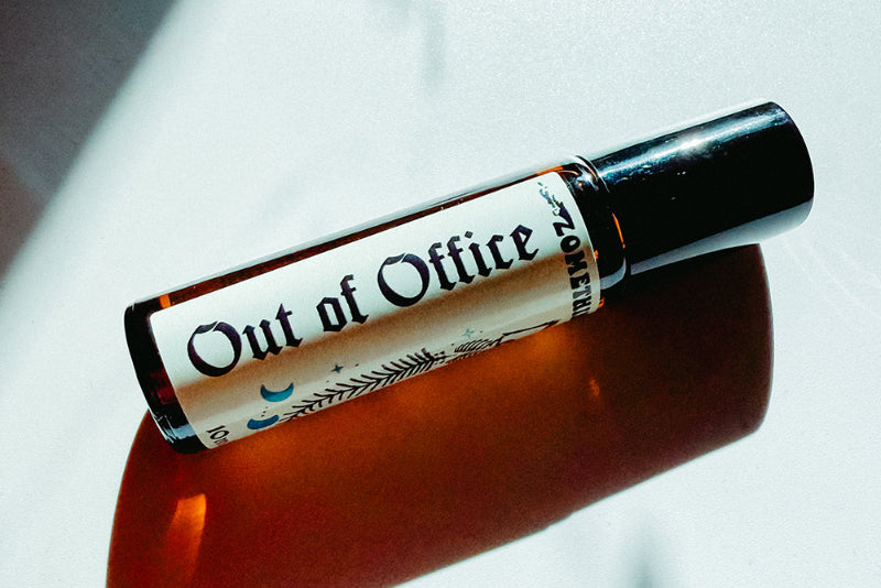 A small amber glass roller bottle with a cream label reading 'Out of Office' and a black cap, photographed diagonally on a glossy white surface with a strong orange reflection.